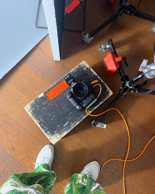 Overhead shot of a black camera on a wooden surface near a light stand and a person wearing white shoes and patterned pants.