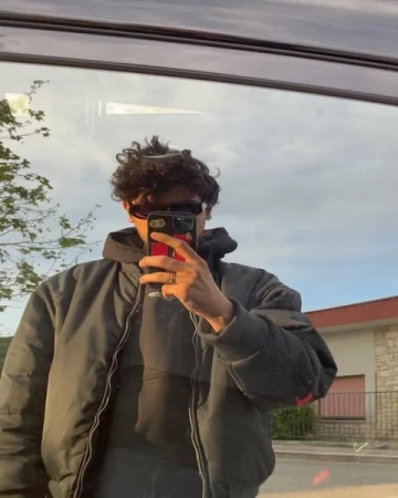Man wearing a dark puffer jacket and sunglasses taking a mirror selfie in a car window reflection in Paris.
