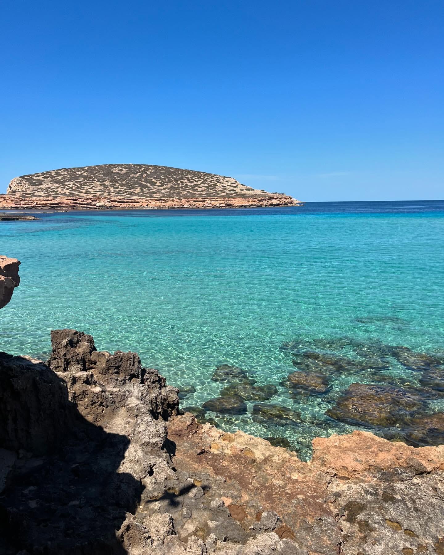Scenic view of clear turquoise water, rocky formations, and a distant island under a bright blue sky on a sunny day.