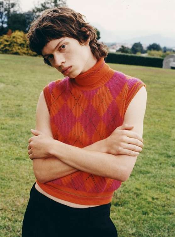 Young adult poses outdoors wearing a pink-and-orange sleeveless argyle turtleneck sweater with black pants on a grass lawn.