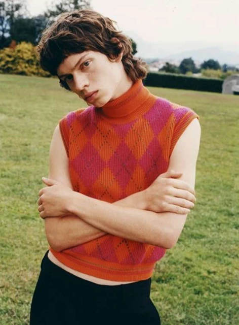 Young adult poses outdoors wearing a pink-and-orange sleeveless argyle turtleneck sweater with black pants on a grass lawn.