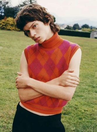 Young adult poses outdoors wearing a pink-and-orange sleeveless argyle turtleneck sweater with black pants on a grass lawn.