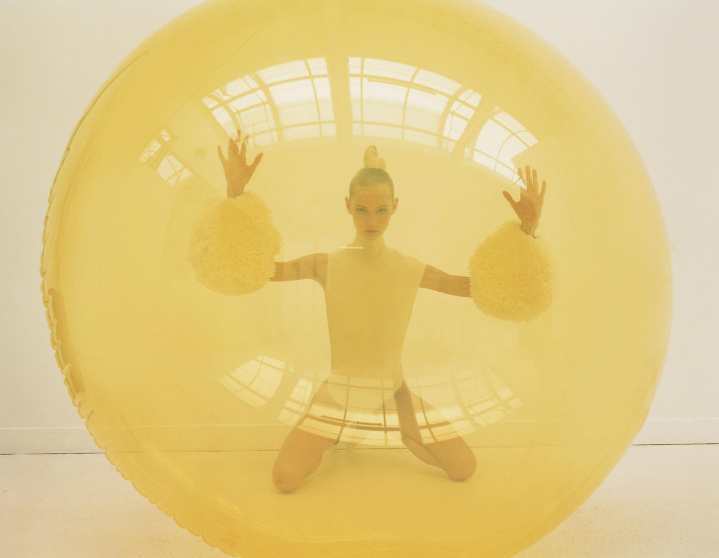 Blonde woman with hands up in a yellow transparent inflatable bubble. The woman wears a yellow top, a skirt, and puffy yellow wristbands.