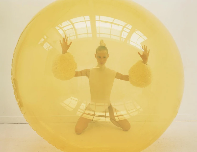 Blonde woman with hands up in a yellow transparent inflatable bubble. The woman wears a yellow top, a skirt, and puffy yellow wristbands.