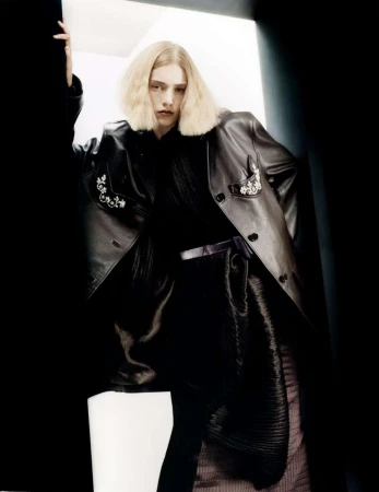 Fashionable adult poses indoors wearing a black leather jacket with decorative embellishments, standing near a dark blue wall.