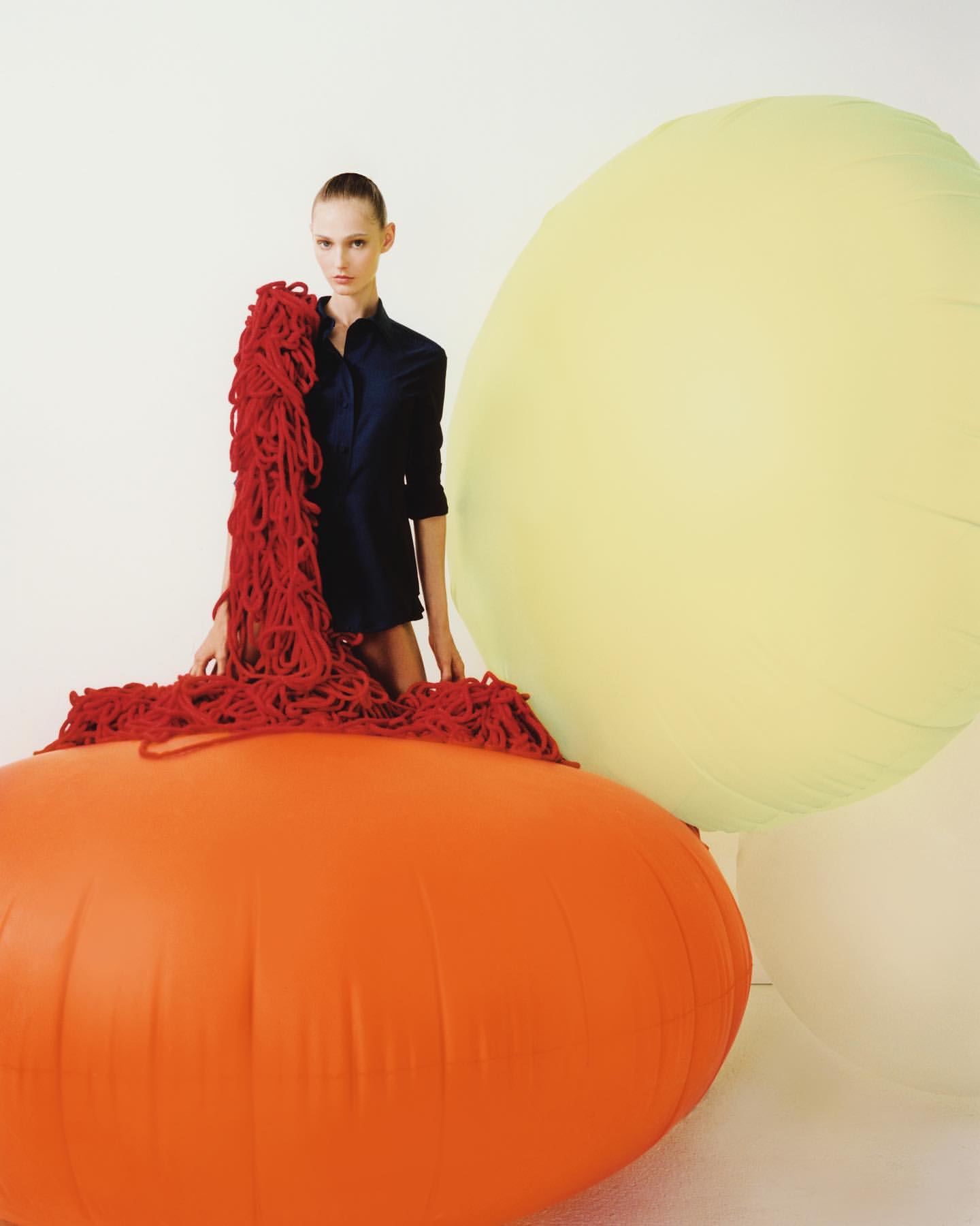 Fashion shot of a model posing with bright oversized balloons, wearing a dark shirt, and red tangled ropes in a brightly lit studio.
