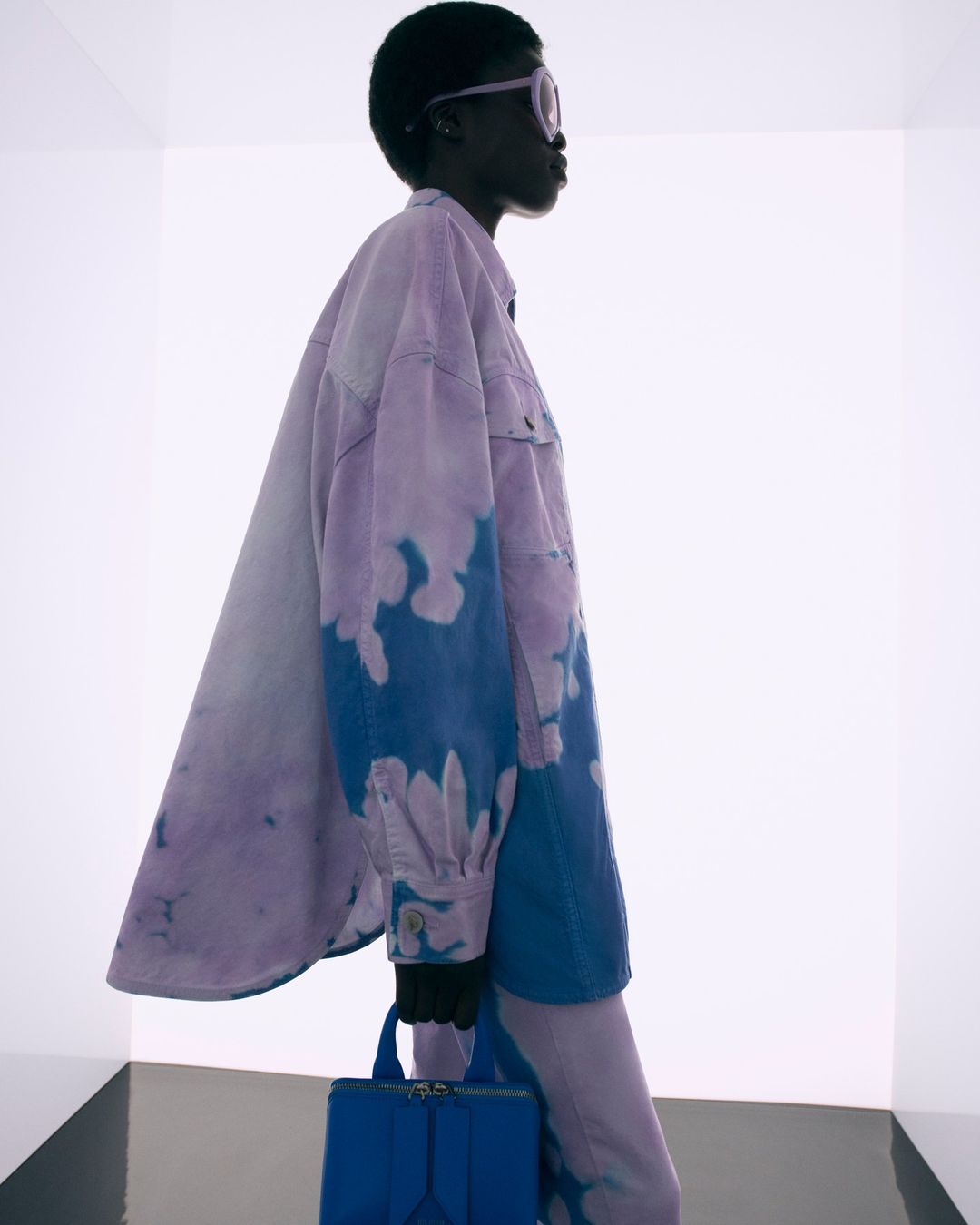 Model in profile wearing a tie-dye purple and blue jacket holding a blue handbag against a white studio backdrop