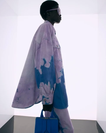 Model in profile wearing a tie-dye purple and blue jacket holding a blue handbag against a white studio backdrop