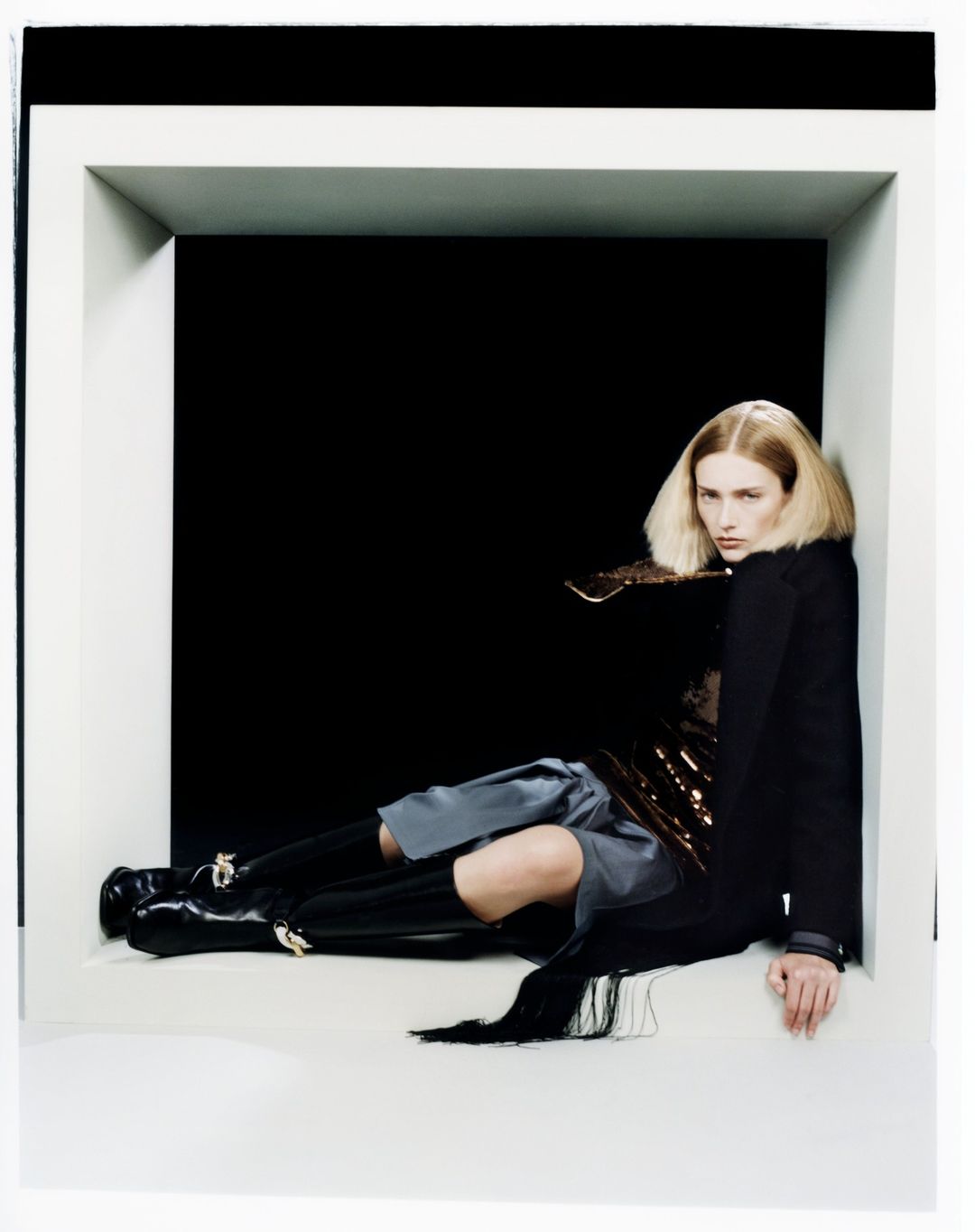 Model with blonde hair reclining inside a large white square architectural frame wearing black coat and boots.
