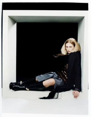 Model with blonde hair reclining inside a large white square architectural frame wearing black coat and boots.