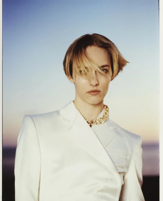Woman in a white suit and statement necklace stands against a light blue and orange sky in a fashion-style portrait. 