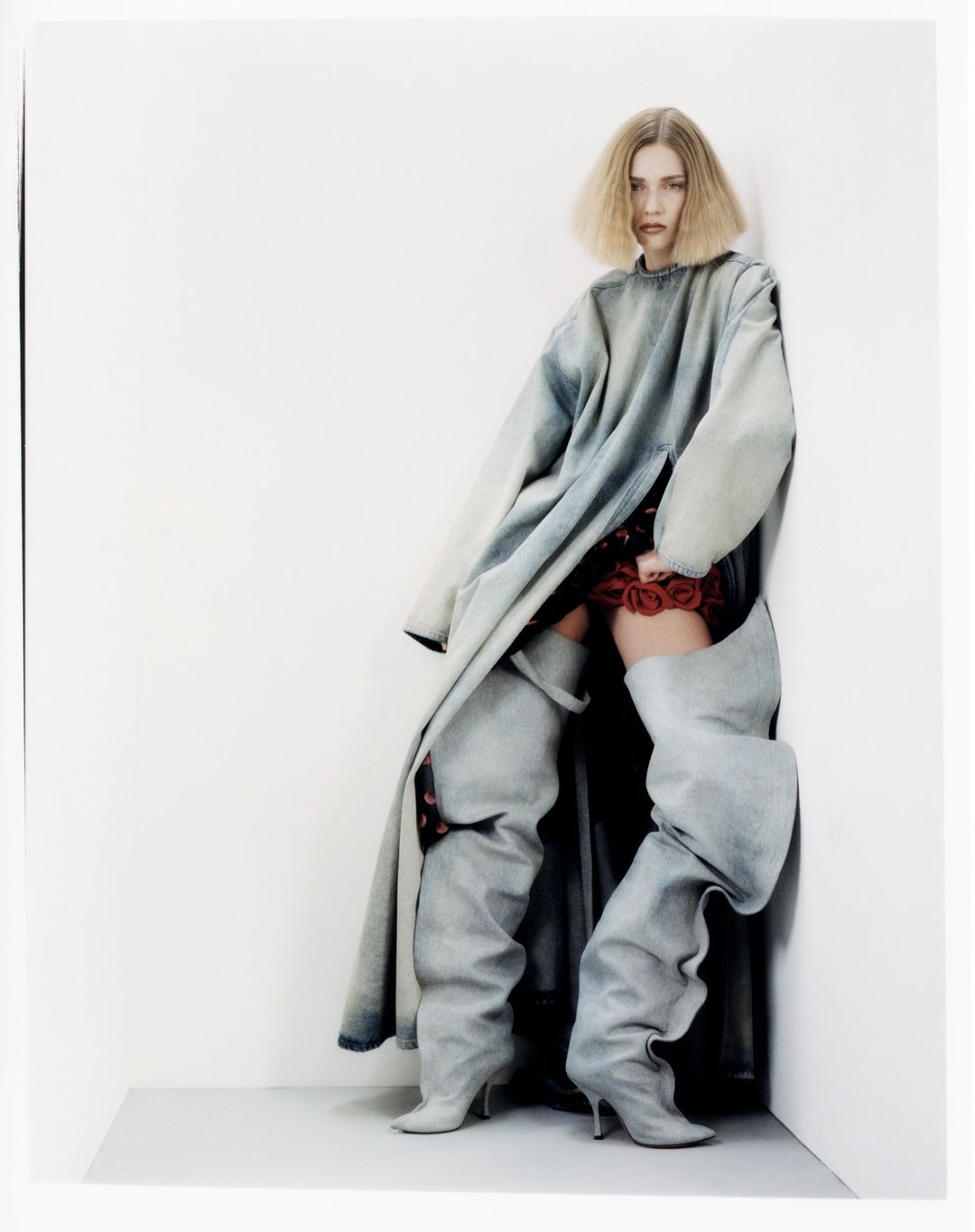 Fashion photo of a blonde woman posing in a light blue oversized denim coat and gray thigh-high denim boots against a white wall.