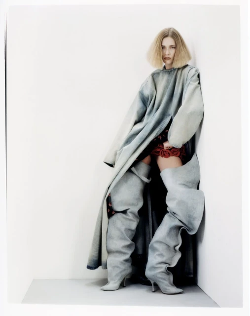 Fashion photo of a blonde woman posing in a light blue oversized denim coat and gray thigh-high denim boots against a white wall.