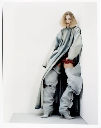 Fashion photo of a blonde woman posing in a light blue oversized denim coat and gray thigh-high denim boots against a white wall.