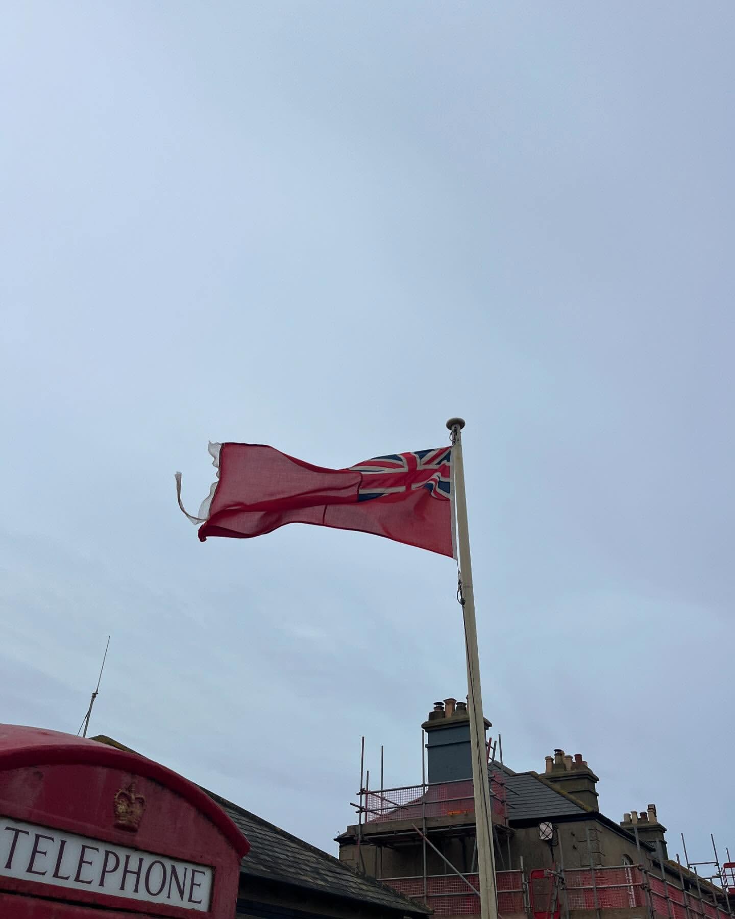 British Red Ensign flag waving in the wind against a cloudy sky, with a telephone box and scaffolding visible in the background.