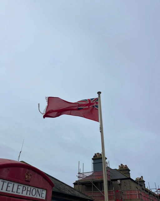 British Red Ensign flag waving in the wind against a cloudy sky, with a telephone box and scaffolding visible in the background.