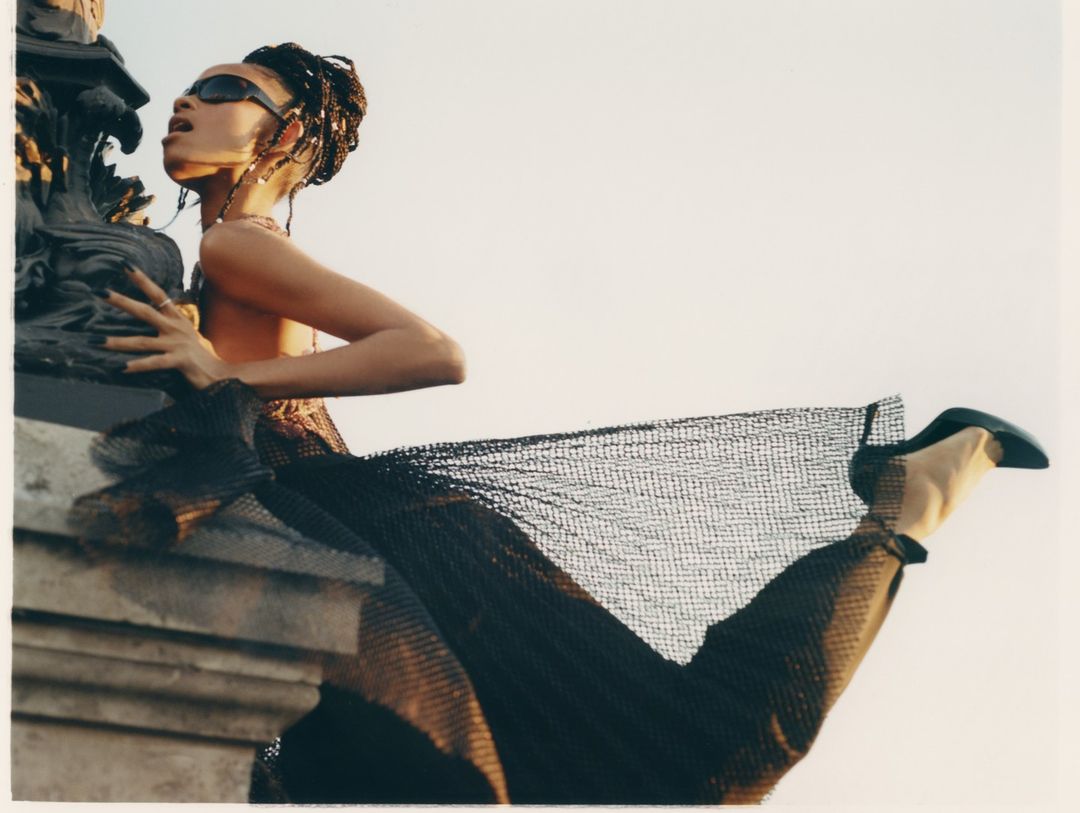 Stylish woman in sunglasses poses with a textured column, wearing a sheer net dress and heeled shoes against a light sky background.