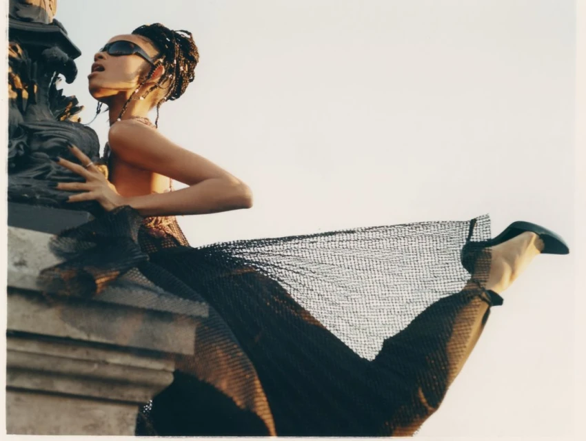 Stylish woman in sunglasses poses with a textured column, wearing a sheer net dress and heeled shoes against a light sky background.