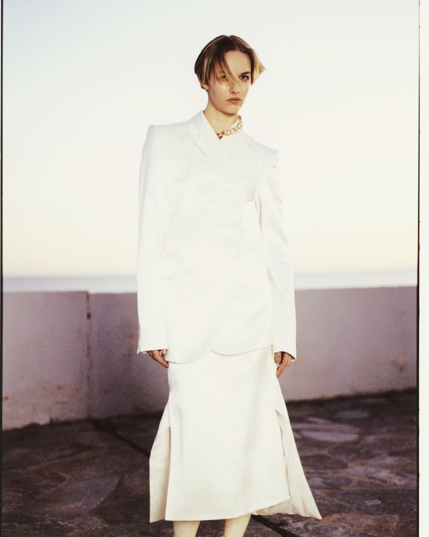 Model wearing a white structured blazer and skirt standing outdoors by a low wall with soft daylight captured by Antonio Dicorato