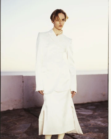 Model wearing a white structured blazer and skirt standing outdoors by a low wall with soft daylight captured by Antonio Dicorato