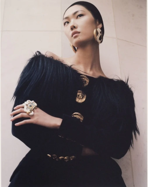 Fashion shot of an Asian woman wearing a black jacket with gold buttons, statement earrings, and an oversized ring, gazing upward in studio setting.