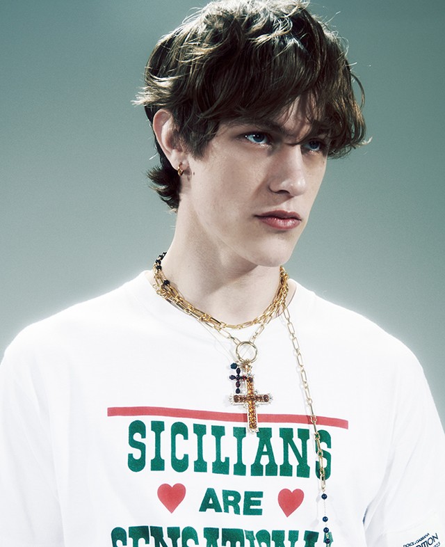 Fashionable young adult with tousled hair wears a branded white tee and gold chains in a studio headshot for a stylish fashion campaign.