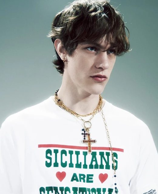Fashionable young adult with tousled hair wears a branded white tee and gold chains in a studio headshot for a stylish fashion campaign.
