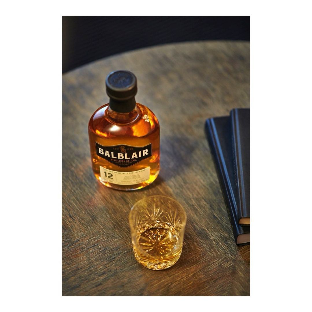 Still life of Balblair 12 whisky bottle and glass on a wooden table beside a navy book, with a modern dark background.