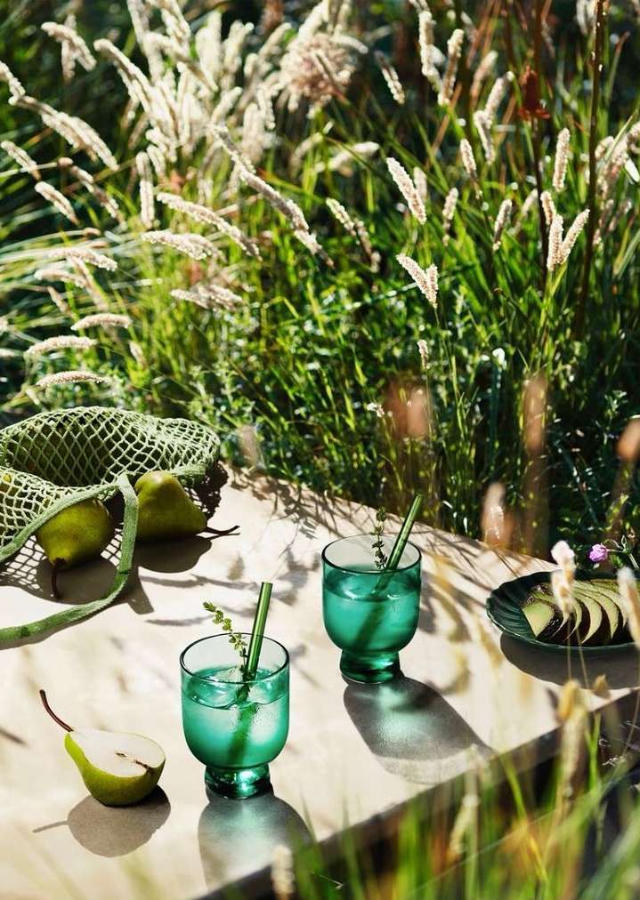 Two emerald-green stemmed glasses with cocktails and green straws on a table, pears and grasses around, outdoors in the sunlight.