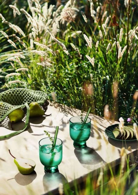 Two emerald-green stemmed glasses with cocktails and green straws on a table, pears and grasses around, outdoors in the sunlight.