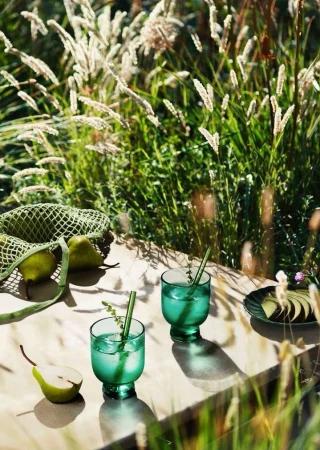 Two emerald-green stemmed glasses with cocktails and green straws on a table, pears and grasses around, outdoors in the sunlight.