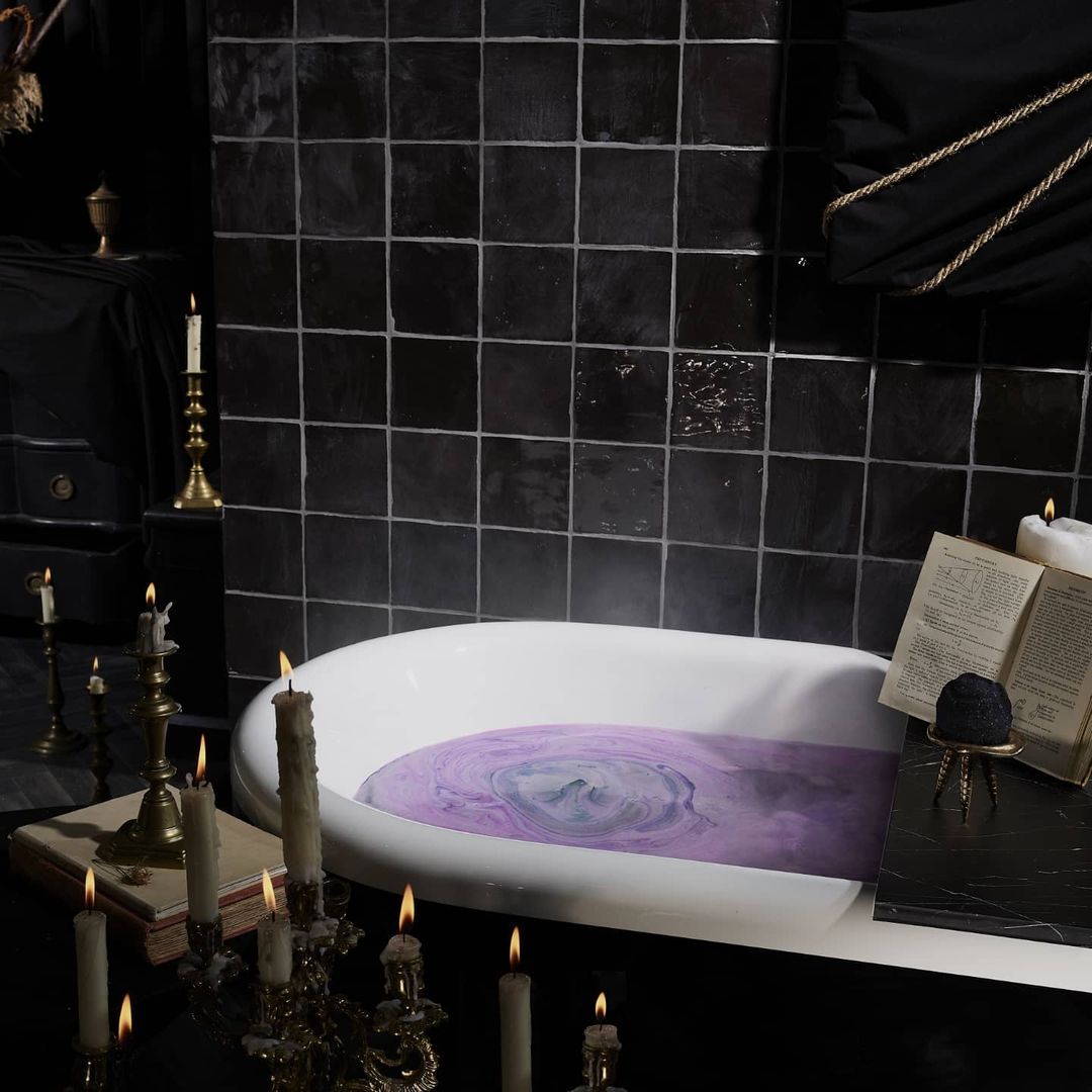A white, claw-footed bathtub filled with swirling purple water sits in a dark, tiled bathroom illuminated by candlelight.
