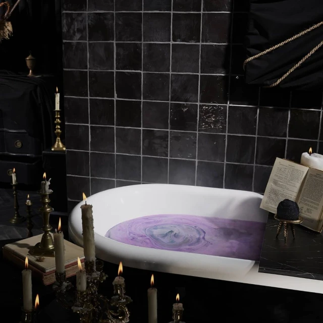 A white, claw-footed bathtub filled with swirling purple water sits in a dark, tiled bathroom illuminated by candlelight.
