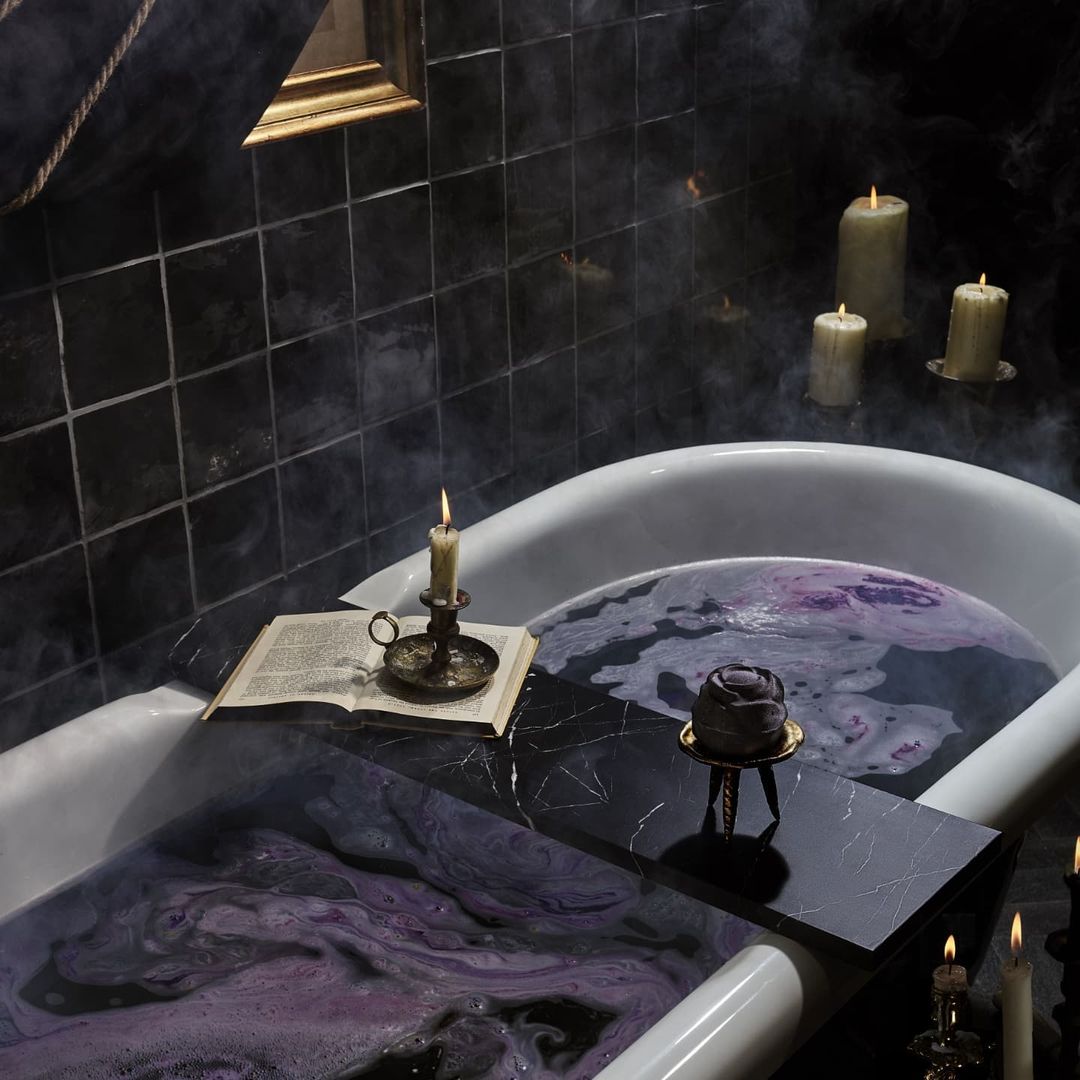 Bathtub filled with purple water, candles, and a book, creating a mystical and relaxing spa atmosphere in a dark-tiled bathroom.