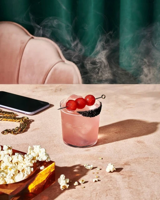 Pink cocktail in a glass with grape garnish, surrounded by popcorn, a phone, and gold chain, set against a green curtain background.