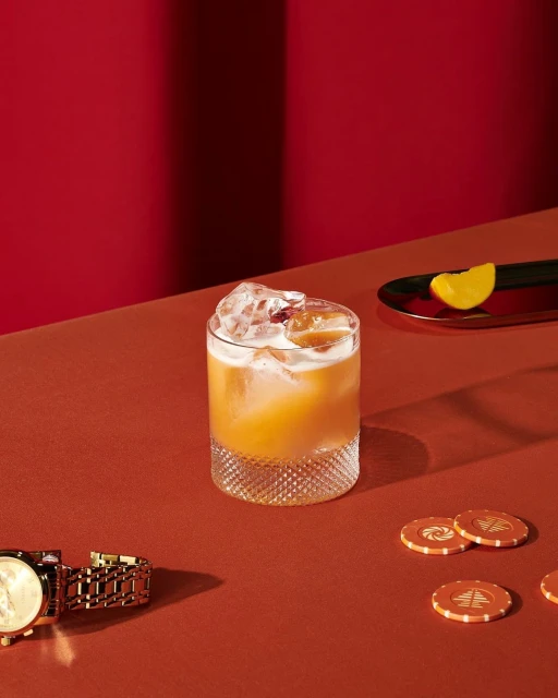 A faceted glass filled with amber liquid and ice, accompanied by a citrus slice, poker chips, and a gold watch on a red surface.