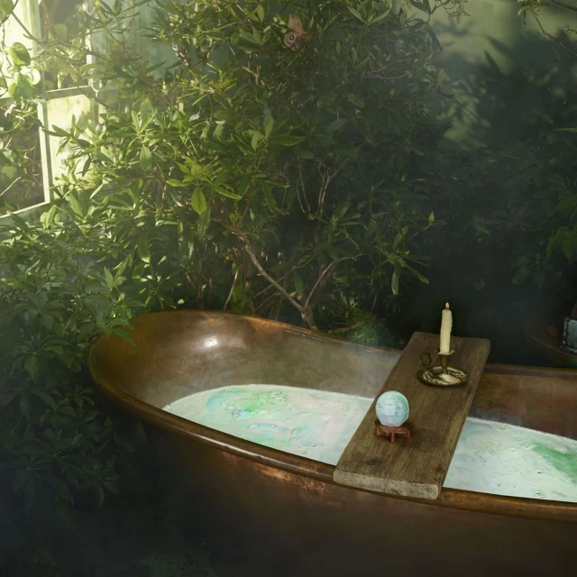 Copper bathtub filled with bubble bath sits on a wooden plank surrounded by green foliage, lit by a single candle.