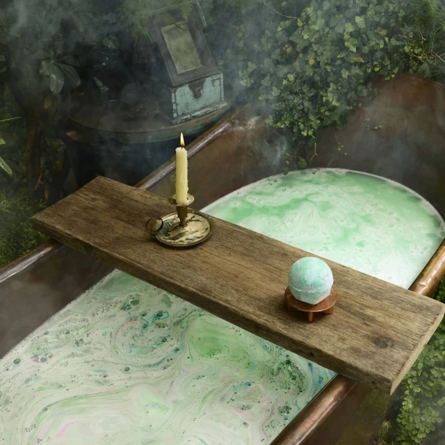 Close-up of a rustic copper bathtub filled with bubbly green water. A wooden plank holds a candle, holder, and bath bomb.