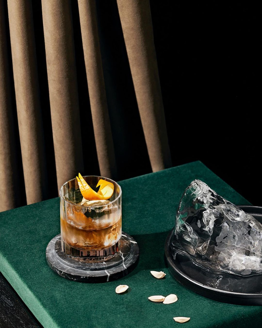 A cocktail in a clear glass with an orange peel garnish is shown alongside a large piece of ice, staged on a green surface.