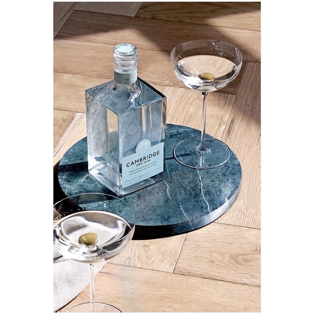 Cambridge Dry Gin bottle and two martini glasses with olives, presented on a round marble tray, sitting on a herringbone wood floor.
