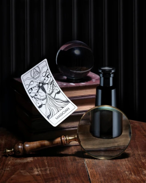 A composition with a crystal ball, tarot card, magnifying glass, books, and black bottle on a wooden table with dark backdrop.