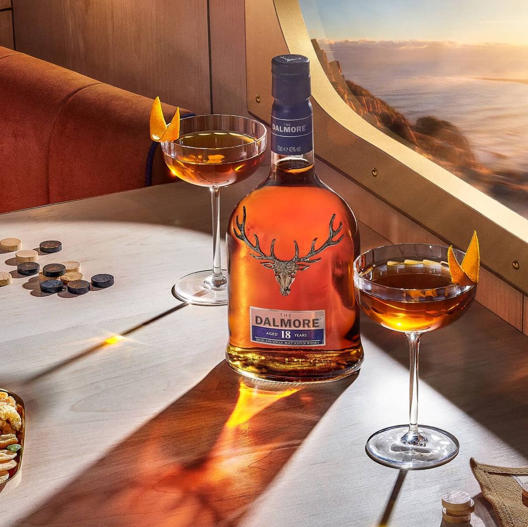 Bottle of Dalmore 18 year scotch whisky on a light wood table with two cocktails and a window view overlooking water and land.