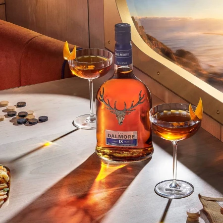 Bottle of Dalmore 18 year scotch whisky on a light wood table with two cocktails and a window view overlooking water and land.
