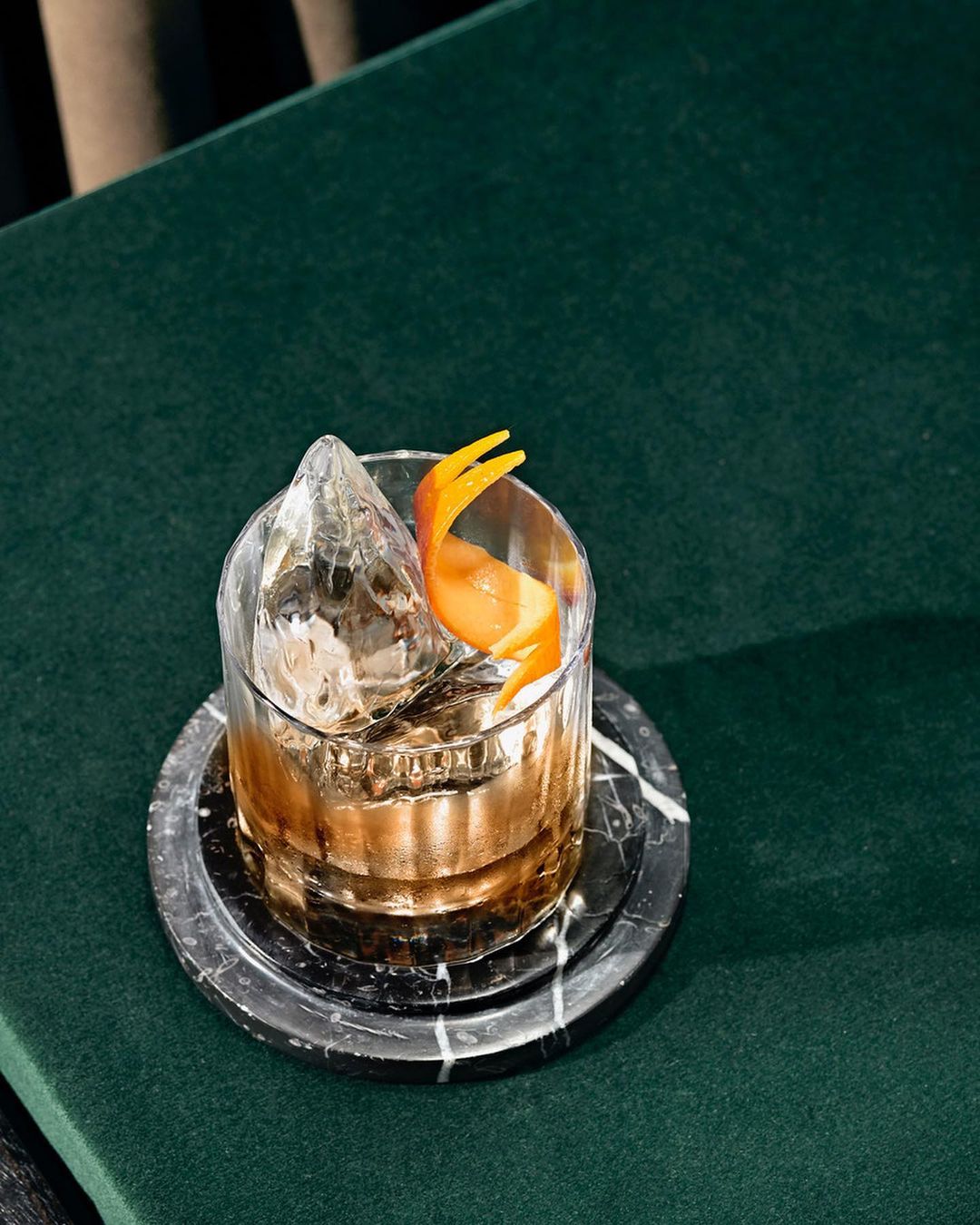 A cocktail in a rocks glass with a large ice cube and orange peel garnish, set on a black marble coaster over green velvet.
