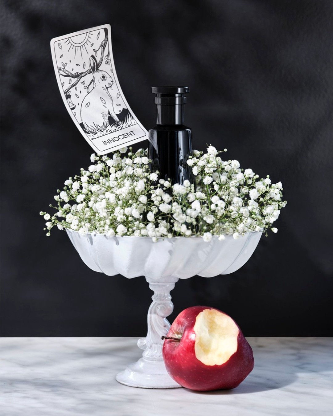 Still life composition: A black bottle surrounded by white flowers in a white pedestal bowl next to a bitten red apple and card.