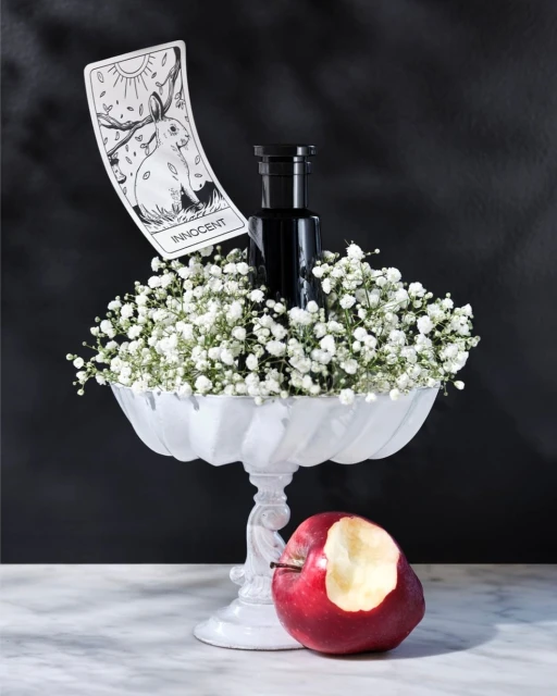 Still life composition: A black bottle surrounded by white flowers in a white pedestal bowl next to a bitten red apple and card.