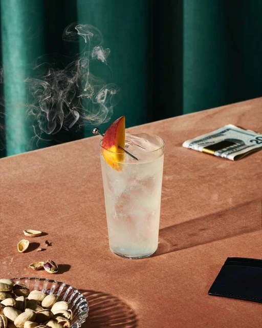 A refreshing cocktail with a peach garnish and pistachio nuts is on a table with smoke, against a green curtain background.