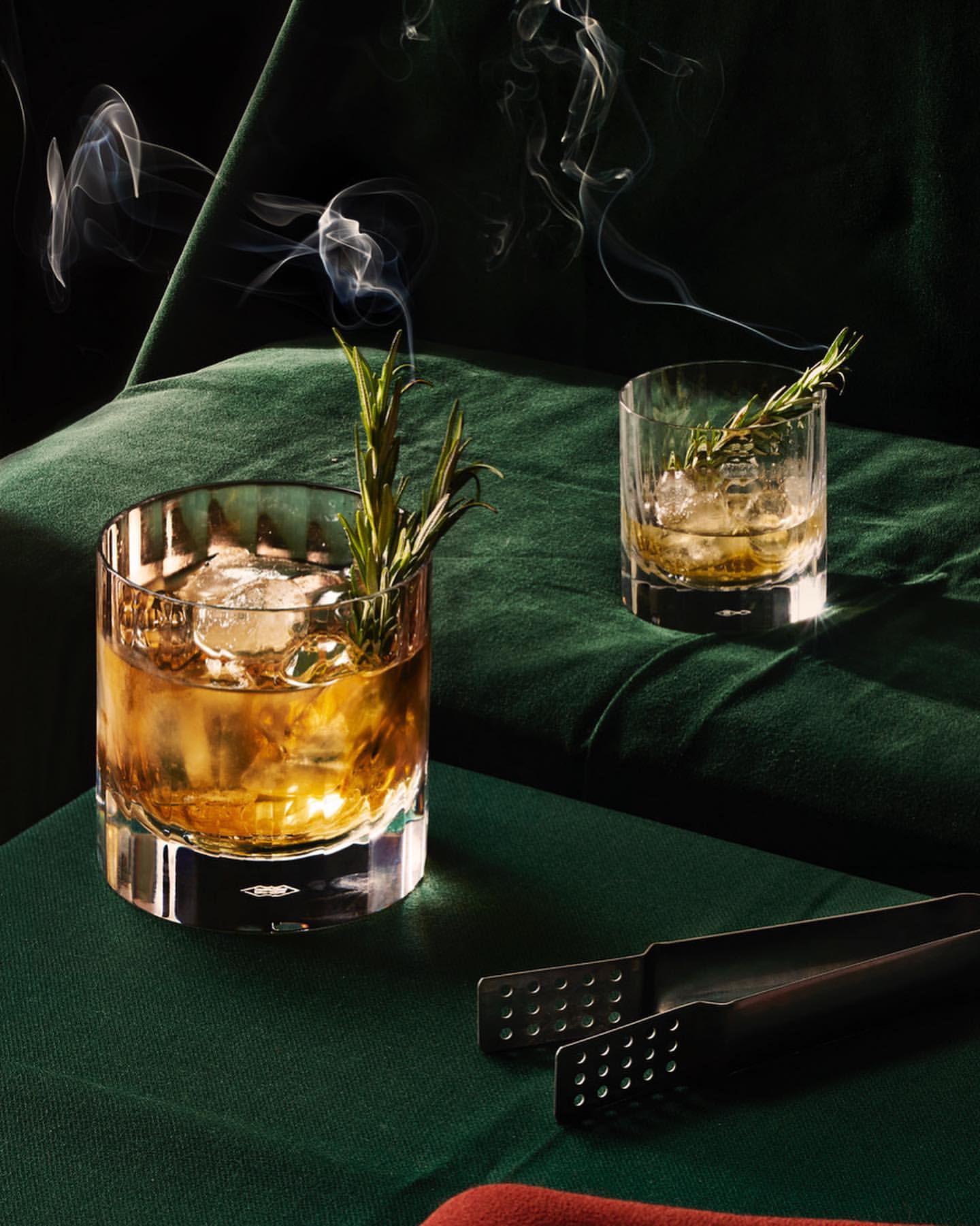 Two glasses of cocktail with ice and rosemary sprigs sit on a green cloth with smoke rising in a dark setting, served with bar tongs.