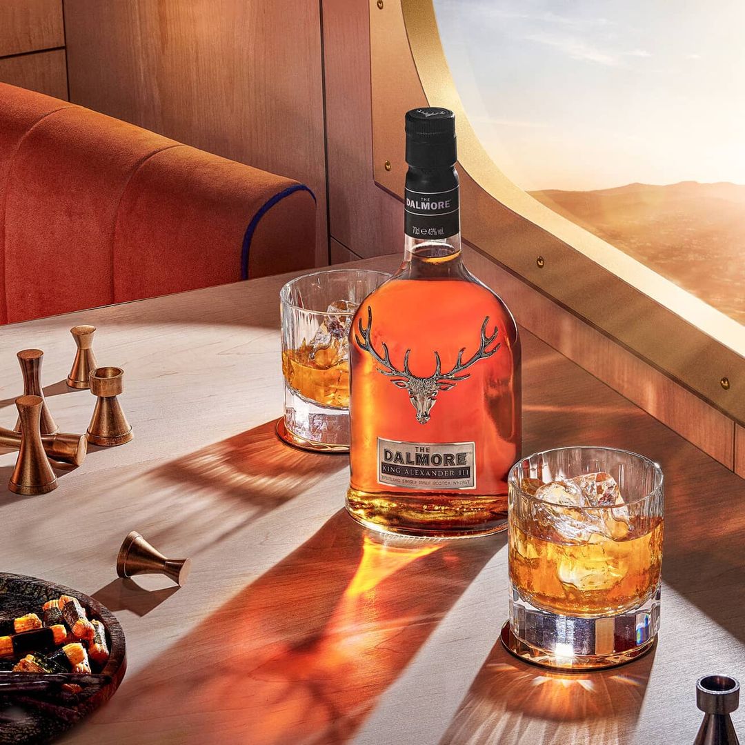 Bottle of Dalmore King Alexander III scotch whiskey on a wooden table near two glasses of scotch and a window showing a desert landscape.
