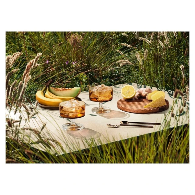 Two amber cocktail glasses, bananas, melon, lemon, and ginger on a white surface outdoors with tall grasses in background.
